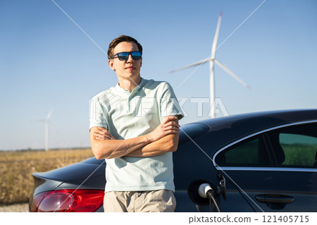 Man with charging electric car. Wind turbines in the background Man with charging electric car. Wind turbines in the background 121405715