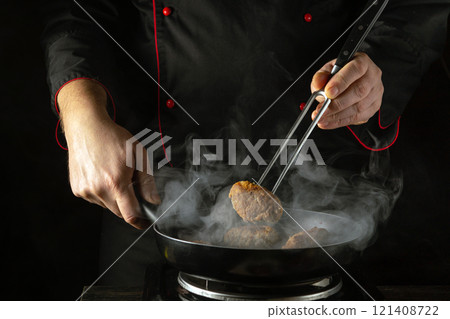 The chef, dressed in a black robe with red accents, expertly holds a fork to sear a cutlet in a frying pan. Steam rises sharply, creating an atmosphere of intense culinary focus The chef, dressed in a black robe with red accents, expertly holds a fork to sear a cutlet in a frying pan. Steam rises sharply, creating an atmosphere of intense culinary focus 121408722