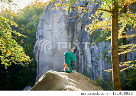 Woman practicing yoga outdoors in forest. Barefoot female on yoga mat surrounded by trees and large rocks, which suggests peaceful, natural environment ideal for meditation or yoga practice. Woman practicing yoga outdoors in forest. Barefoot female on yoga mat surrounded by trees and large rocks, which suggests peaceful, natural environment ideal for meditation or yoga practice. 121408926