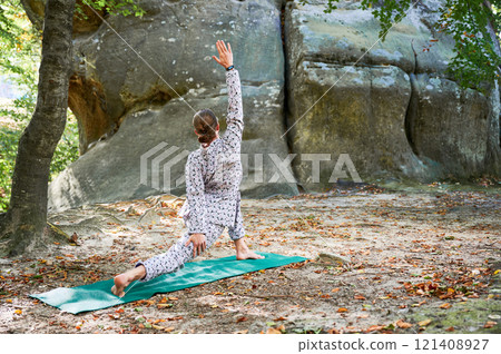 Woman practicing yoga outdoors in forest. Barefoot female on yoga mat surrounded by trees and large rocks, which suggests peaceful, natural environment ideal for meditation or yoga practice. 121408927