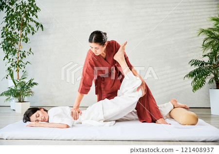 Masseuse making thai yoga massage. Woman therapist doing traditional massage treatment, stretching female patient's body. Client lying on mat, enjoying hand massage in calm atmosphere. 121408937