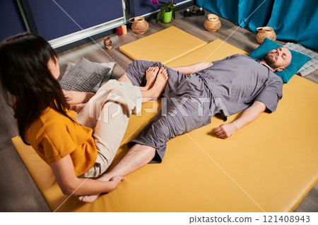 Masseuse making thai yoga massage. Woman therapist doing traditional massage treatment, stretching male patient's body. Client lying on mat, enjoying hand massage in calm atmosphere. 121408943