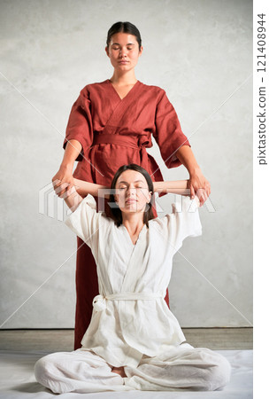 Masseuse making thai yoga massage. Woman therapist doing traditional massage treatment, stretching female patient's body. Client sitting on mat, enjoying hand massage in calm atmosphere. Masseuse making thai yoga massage. Woman therapist doing traditional massage treatment, stretching female patient's body. Client sitting on mat, enjoying hand massage in calm atmosphere. 121408944