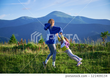 Young woman tourist swings, spending time together with little daughter on grassy hill, set against stunning mountain backdrop. Joyful moment captures essence of carefree fun and natural beauty. Young woman tourist swings, spending time together with little daughter on grassy hill, set against stunning mountain backdrop. Joyful moment captures essence of carefree fun and natural beauty. 121409134