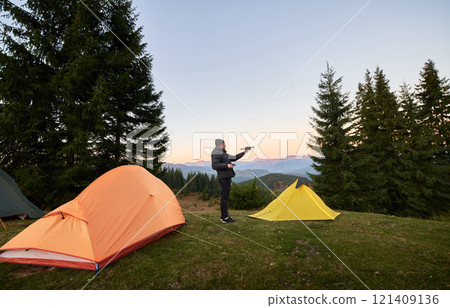 Male traveler standing on grassy hill near camp tents and holding quadcopter for aerial photography, capturing picturesque landscape. Sky softly lit with pastel colors from early morning light. 121409136