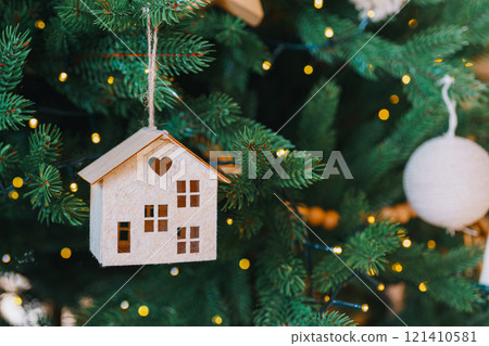 Handcrafted wooden house ornament hanging on Christmas tree branch with warm lights background 121410581