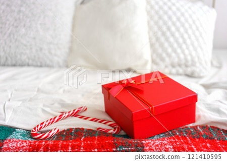Bright red gift box placed on a cozy white bed with a festive blanket during the holiday season 121410595