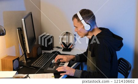 In a cozy home studio, a man deeply focuses on composing a music track using a MIDI keyboard controller, wearing headphones and surrounded by monitors and musical equipment In a cozy home studio, a man deeply focuses on composing a music track using a MIDI keyboard controller, wearing headphones and surrounded by monitors and musical equipment 121410602