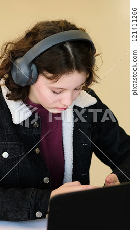 A young girl with headphones concentrates on writing in her notebook while seated at a desk. This image highlights the importance of focused study and multitasking in modern educational practices. 121411266