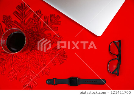 A transparent mug of coffee, in which foam and a bottle are visible, stands on a red napkin in the form of a snowflake on a red background, next to it are a gray laptop, glasses in a transparent frame A transparent mug of coffee, in which foam and a bottle are visible, stands on a red napkin in the form of a snowflake on a red background, next to it are a gray laptop, glasses in a transparent frame 121411700