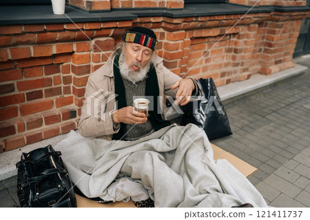 Happy elderly homeless man warming up with cup of coffee on cold urban street, evoking sadness and highlighting issue of homelessness in America. Concept of raw reality of poverty and despair. Happy elderly homeless man warming up with cup of coffee on cold urban street, evoking sadness and highlighting issue of homelessness in America. Concept of raw reality of poverty and despair. 121411737