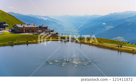 Visitors enjoy the serene atmosphere at Wildkogel water reservoir, surrounded by stunning mountain views of the Austrian Alps, with a fountain enhancing the picturesque landscape. Visitors enjoy the serene atmosphere at Wildkogel water reservoir, surrounded by stunning mountain views of the Austrian Alps, with a fountain enhancing the picturesque landscape. 121411872
