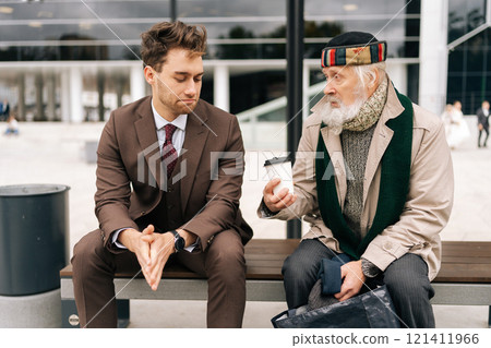 Handsome kindness businessman in formal attire attentively engages in conversation with senior homeless man holding disposable coffee cup, while seated on bench in public area, on cloudy day. 121411966