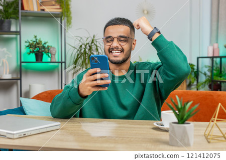 Indian Arabian man at home table use smartphone celebrating success victory winning play online game 121412075