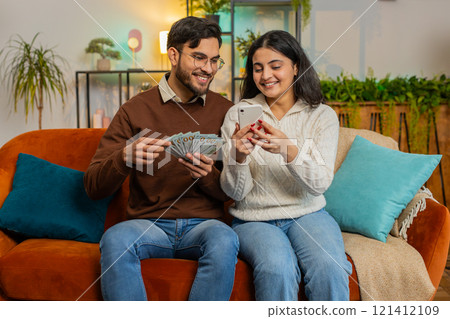 Happy young couple holding money cash and smartphone planning discussing investment sitting on sofa Happy young couple holding money cash and smartphone planning discussing investment sitting on sofa 121412109