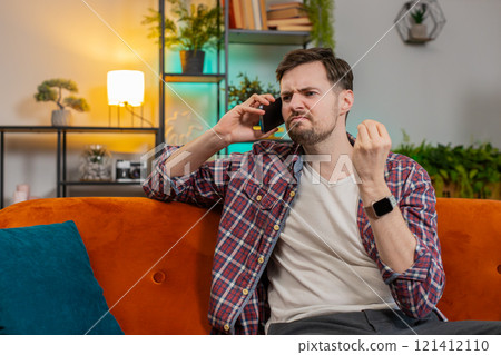 Angry young man arguing talking loudly shouting on smartphone quarrel sitting on couch at home 121412110