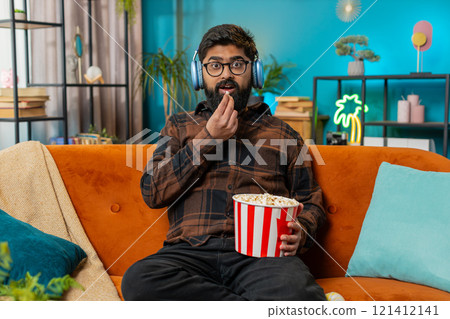 Relaxed Indian man eating popcorn watching movie enjoying film during weekend sitting on home sofa Relaxed Indian man eating popcorn watching movie enjoying film during weekend sitting on home sofa 121412141