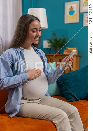 Smiling pregnant woman using smartphone making online purchases for baby sitting on home couch Smiling pregnant woman using smartphone making online purchases for baby sitting on home couch 121412159