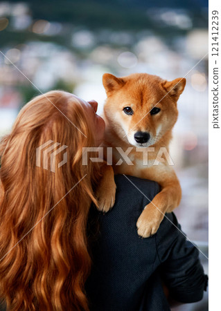A woman with long red hair holds a Shiba Inu dog against a blurred landscape background.  121412239