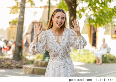 Woman in dress smiling friendly at camera waving hands gesturing hi greeting goodbye on city street 121412338