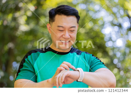 Asian runner man jogging in city park and looking at smartwatch using smart bracelet checking result 121412344
