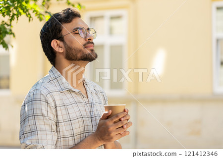 Happy Indian young man enjoying morning coffee hot drink and smiling standing on city street 121412346