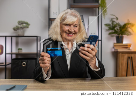 Happy Caucasian senior woman using credit card and smartphone for online shopping payments at office 121412358