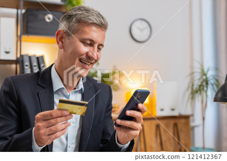 Smiling Caucasian freelancer in formal suit using credit card and smartphone at home office desk Smiling Caucasian freelancer in formal suit using credit card and smartphone at home office desk 121412367