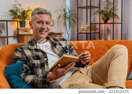 Man reading interesting book turning pages smiling enjoying literature taking a rest on home couch Man reading interesting book turning pages smiling enjoying literature taking a rest on home couch 121412375