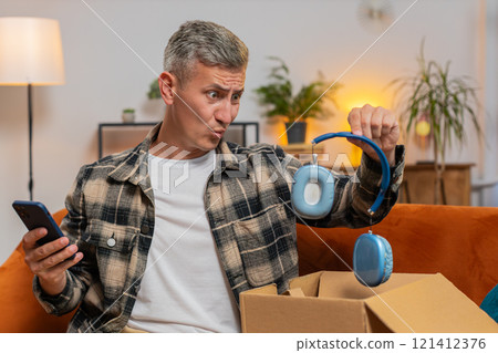Unhappy man unpacking carton box feeling dissatisfied with getting damaged wireless headphones home Unhappy man unpacking carton box feeling dissatisfied with getting damaged wireless headphones home 121412376
