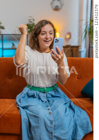 Happy young woman sitting on sofa using smartphone celebrating success victory, winning lottery 121412379