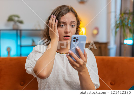 Upset displeased Caucasian woman using smartphone reading bad news sitting on sofa in living room 121412380