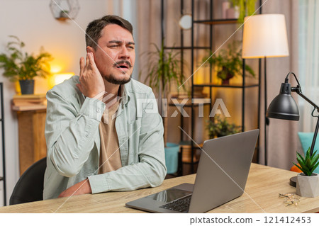 Sick young Caucasian man freelancer sitting at home office table feeling ear pain in living room 121412453
