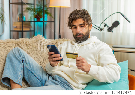 Happy young Hispanic man guy using credit bank card and cellphone while shopping online at home Happy young Hispanic man guy using credit bank card and cellphone while shopping online at home 121412474