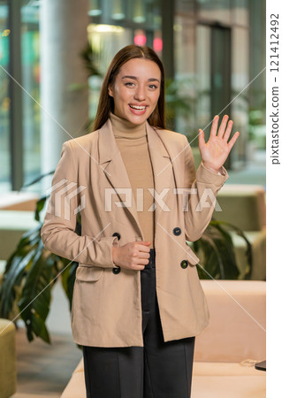 Happy young Caucasian businesswoman looking at camera and waves hand hello in office hotel lobby Happy young Caucasian businesswoman looking at camera and waves hand hello in office hotel lobby 121412492