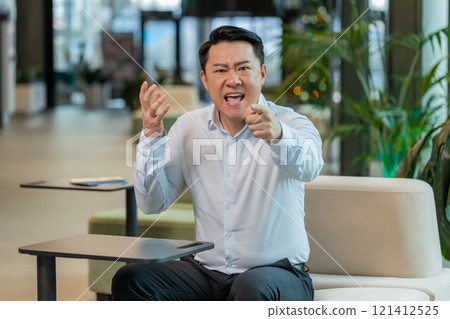 Angry mature boss arguing over video call looking at camera sitting at modern workplace in office Angry mature boss arguing over video call looking at camera sitting at modern workplace in office 121412525