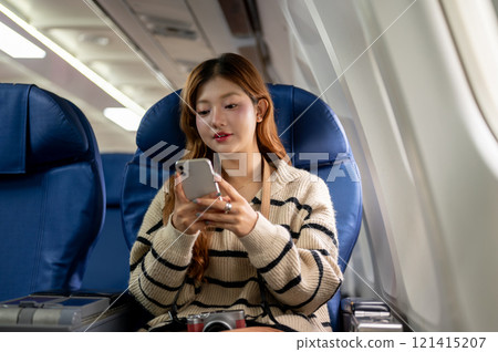 A happy Asian woman in a cozy sweater uses her smartphone during the flight, traveling to somewhere. 121415207