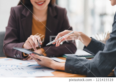 Two businesswomen discussing financial data using tablet and charts at meeting. 121415519