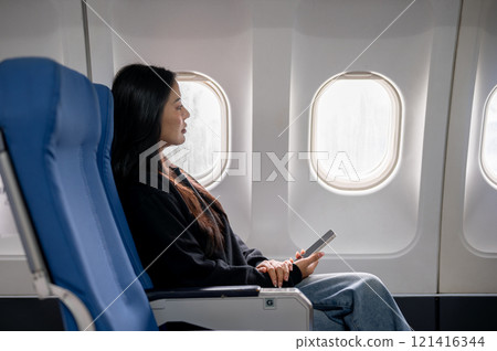 Relaxed Asian female passenger sits at a window seat, looking at the view outside during the flight Relaxed Asian female passenger sits at a window seat, looking at the view outside during the flight 121416344