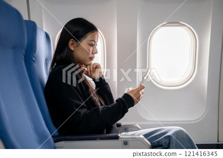 A side view of a relaxed Asian female passenger reading messages on her smartphone during the flight 121416345