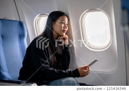 Relaxed Asian female passenger sits at a window seat, looking at the view outside during the flight 121416346