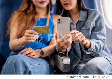 A close-up image of two Asian female friends traveling by plane together, one holding a credit card. 121416376
