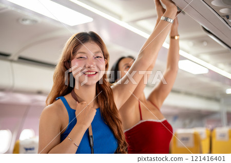 Two Asian girls place their bags in the overhead locker on the plane, with one smiling at the camera 121416401