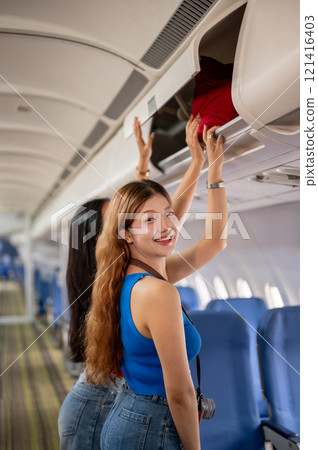 Two Asian girls place their bags in the overhead locker on the plane, with one smiling at the camera 121416403