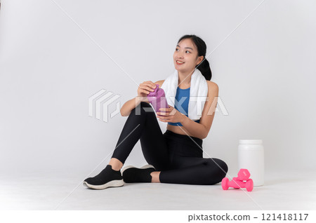 Healthy Asian woman in sportswear sits on a white studio background, holding a protein shake bottle. 121418117