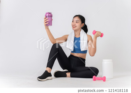 A woman in sportswear sits on a studio background, holding a dumbbell and a protein shake bottle. A woman in sportswear sits on a studio background, holding a dumbbell and a protein shake bottle. 121418118