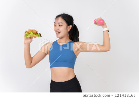 A confident Asian woman in sportswear holding a burger in one hand and a dumbbell in the other. A confident Asian woman in sportswear holding a burger in one hand and a dumbbell in the other. 121418165