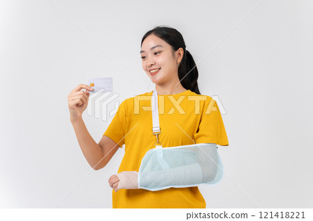A carefree Asian woman with her broken arm, wrapped in a plaster bandage, holding her credit card. 121418221