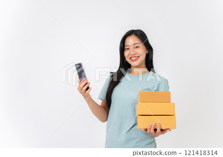 A smiling woman standing against a white studio background, holding cardboard boxes and a smartphone 121418314