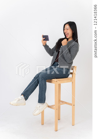 A cheerful Asian woman enjoys watching a sport match or playing a mobile game on her smartphone. 121418366